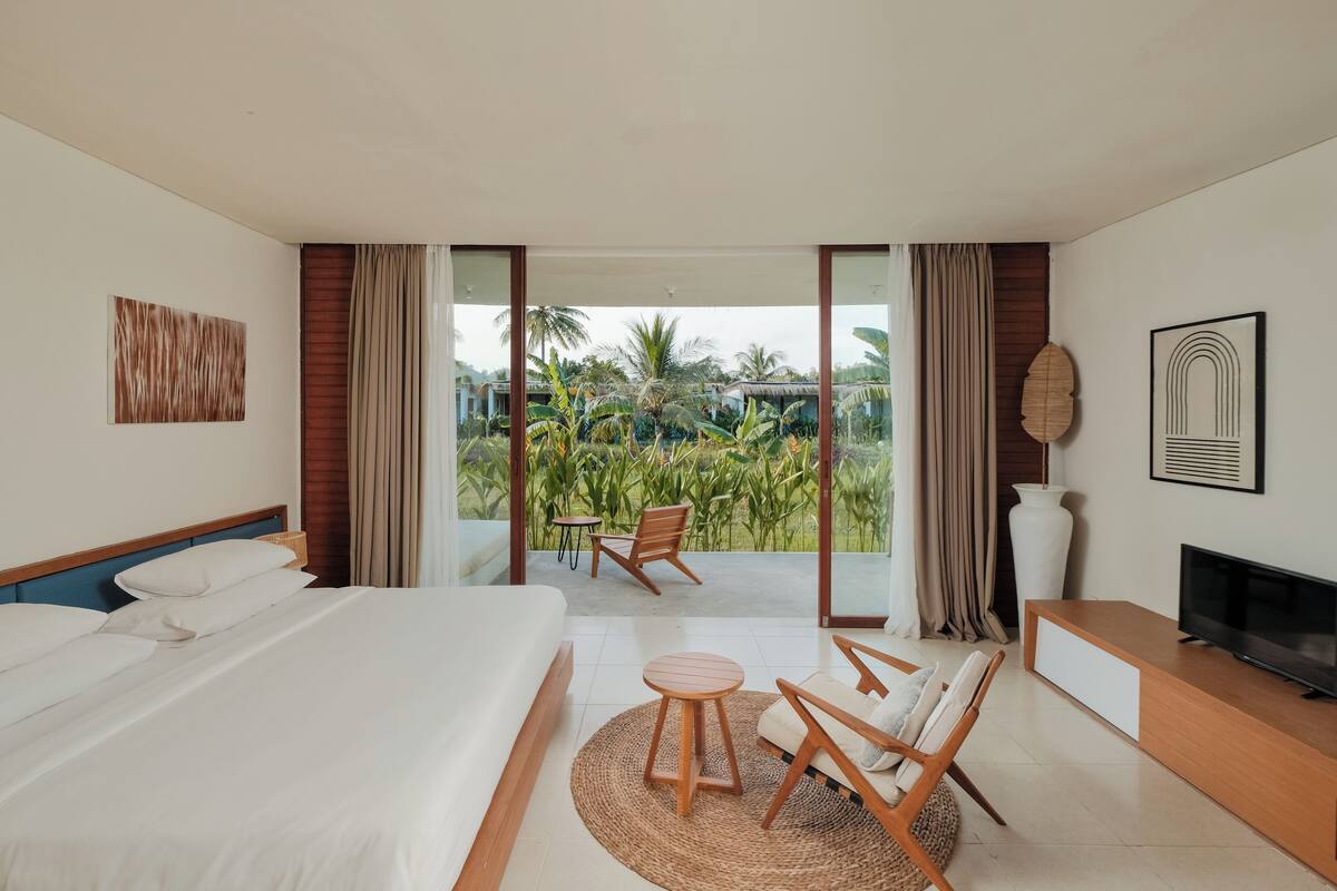 A hotel room with a large bed, a TV, a seating area with a table and chairs, and a balcony with a view of greenery.