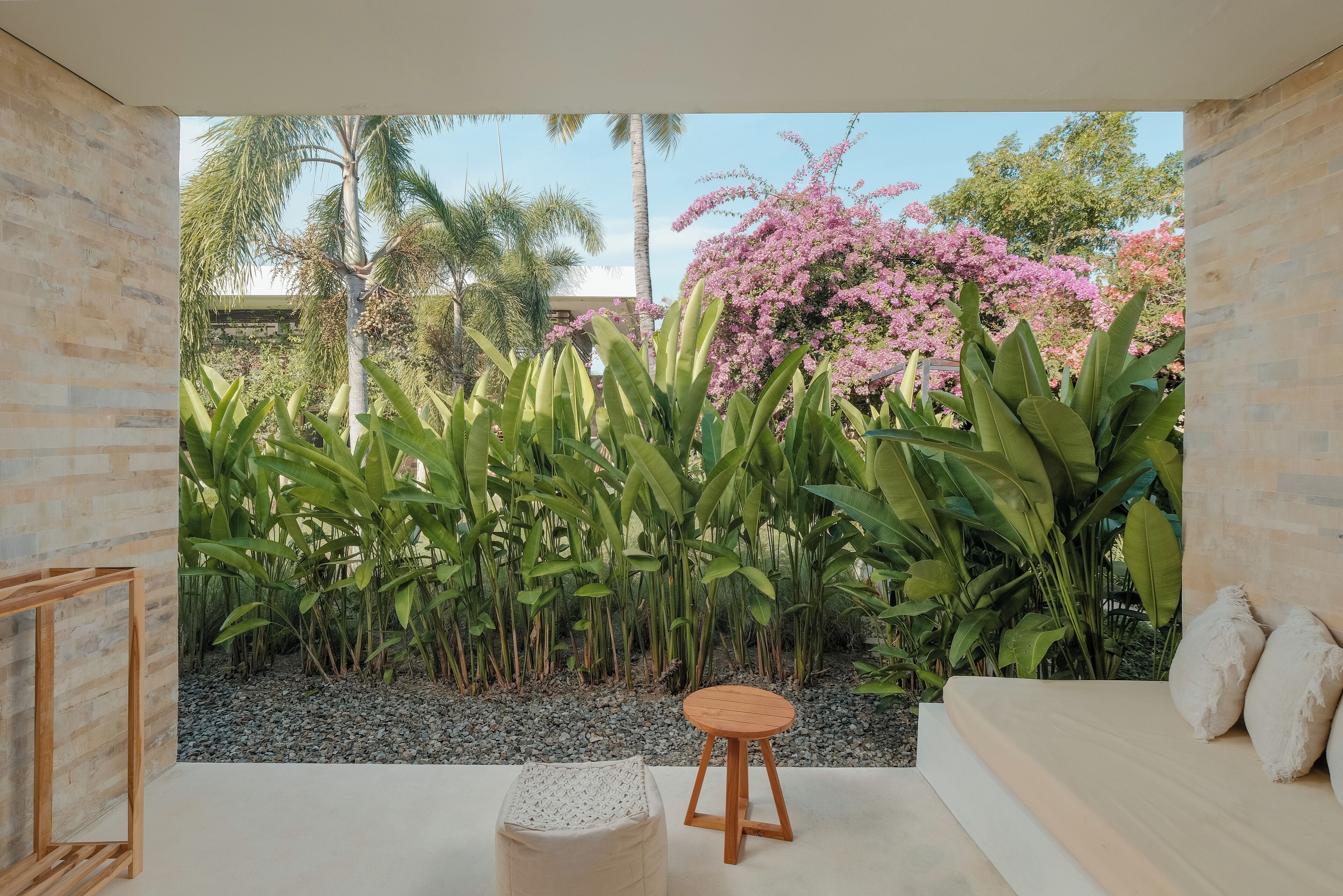 Una habitación con vistas a un jardín, un taburete de madera, un otomano blanco y un sofá con cojines.