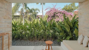 Una habitación con vistas a un jardín, un taburete de madera, un otomano blanco y un sofá con cojines.