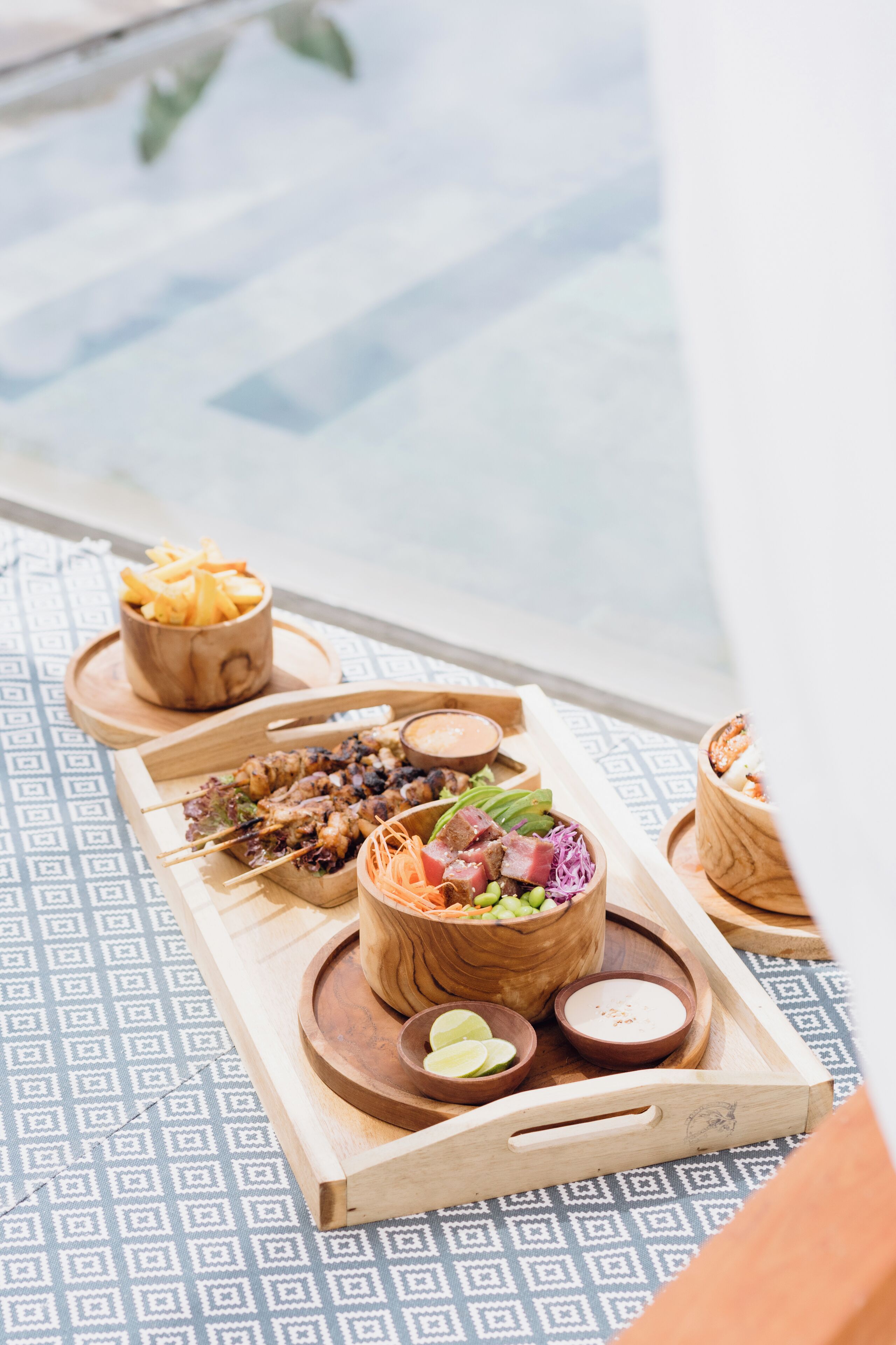 Ein Holztablett mit verschiedenen Speisen, darunter Spieße, ein Salat und Dips, auf einer gemusterten Tischdecke in der Nähe eines Schwimmbeckens.
