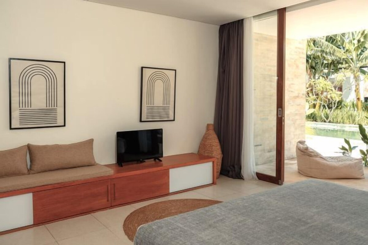 A bedroom with a bed, a TV on a wooden console, a large window with a view of greenery, and a sliding door leading to an outdoor area.