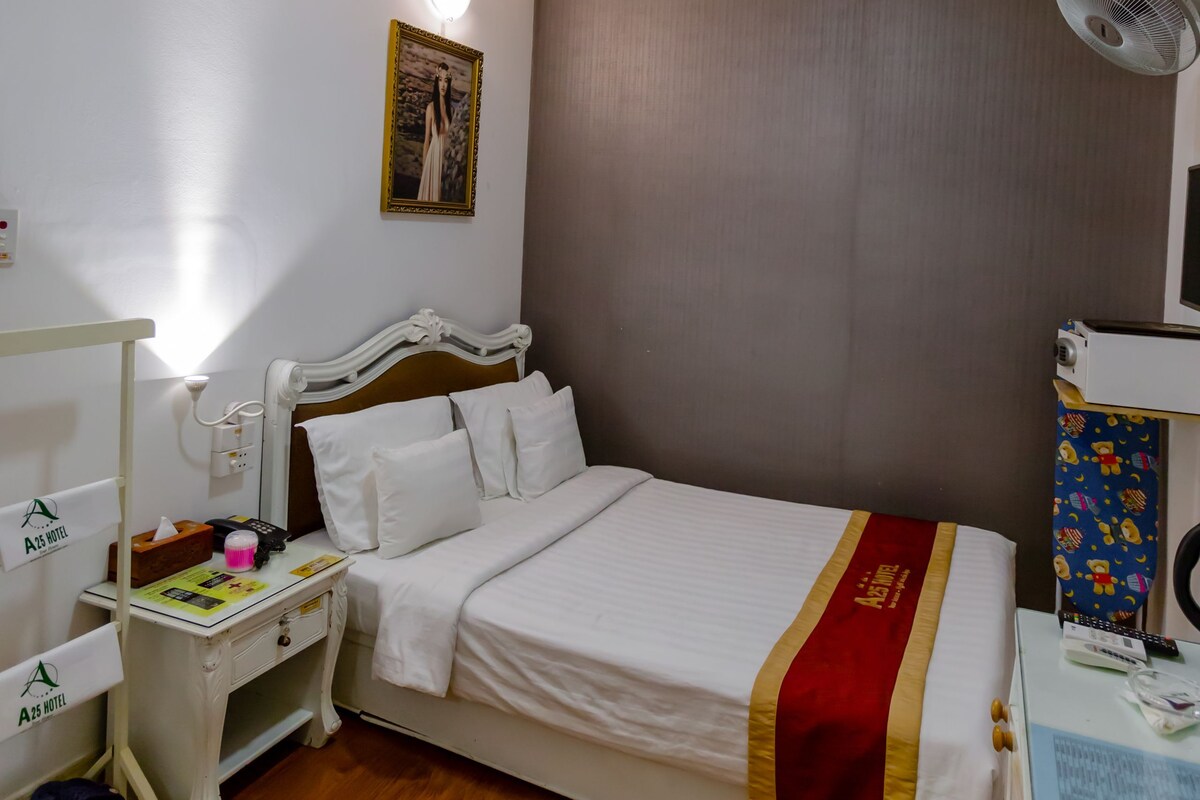 A hotel room with a bed, bedside table, nightstand, and a small desk with a fan.
