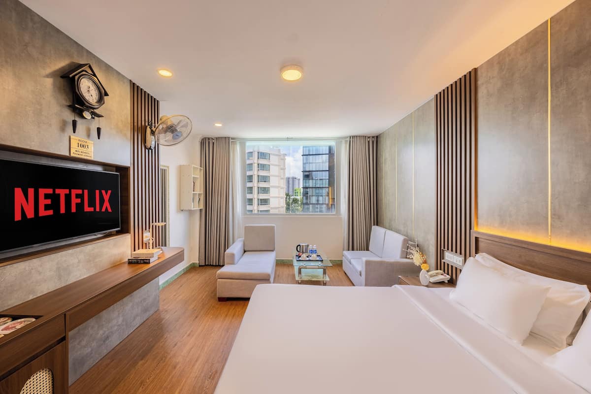 Une chambre d’hôtel avec un grand lit, un téléviseur affichant le logo « NETFLIX », un bureau avec un ventilateur et une vue sur les bâtiments par la fenêtre.