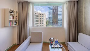 Hotellihuone, jossa on sohva, pieni pöytä, jolla on tarjotin täynnä aamiaistarvikkeita, kirjahylly ja suuri ikkuna, jossa on verhot.