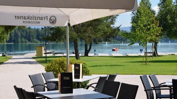 An outdoor seating area with tables and chairs, overlooking a lake with boats and a dock. - Strandhotel Faak (Finkenstein am Faaker See)