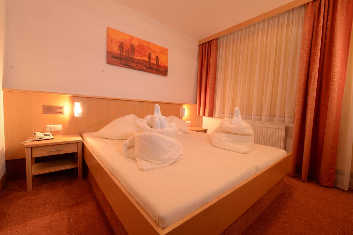 A hotel room with a large bed, two bedside tables, a nightstand, a painting on the wall, and a window with curtains.