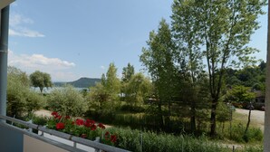 A balcony with a view of a garden, trees, and a distant lake. - Strandhotel Faak (Finkenstein am Faaker See)