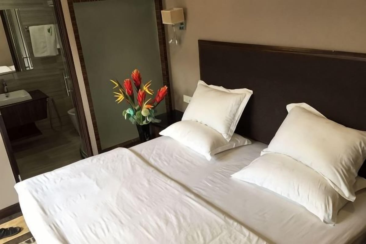 A hotel room with a bed, pillows, a nightstand, a vase with flowers, a mirror, and a bathroom visible through a glass door.