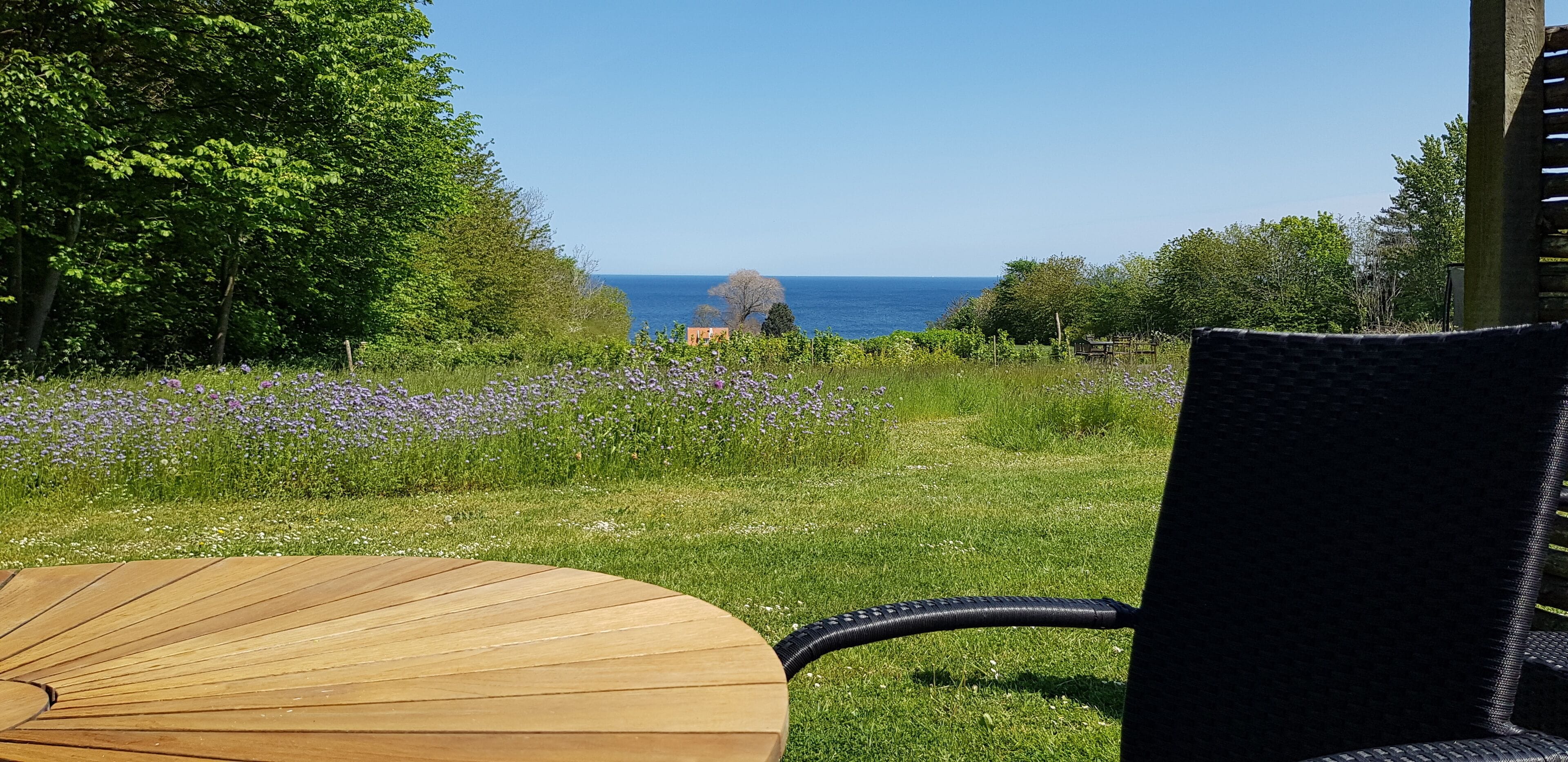 A patio with a round wooden table and a black wicker chair, overlooking a grassy area with purple flowers and a view of the sea.