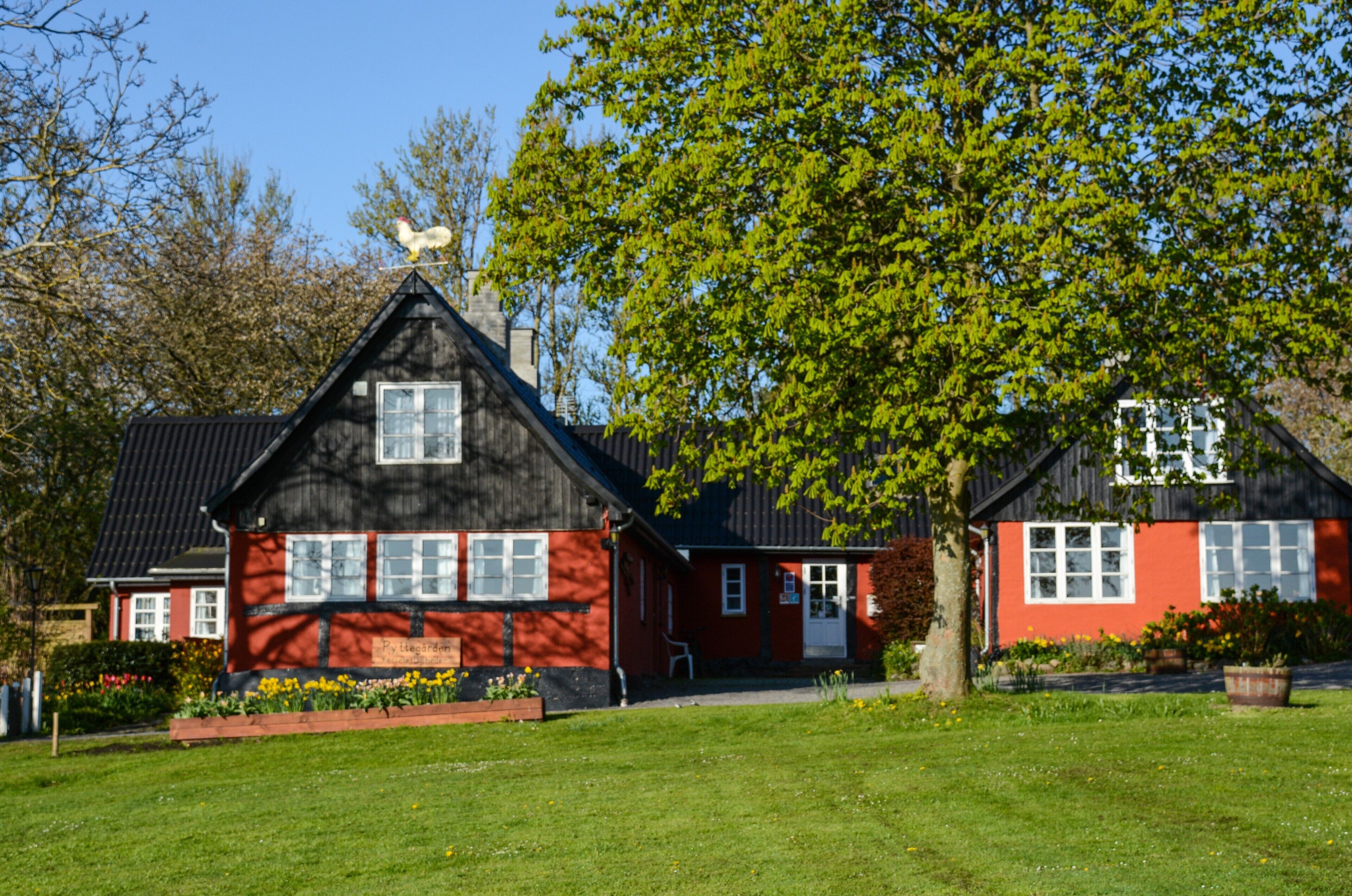 Et traditionelt hus i rødt og sort med skorsten, omgivet af en velplejet græsplæne og blomstrende planter.