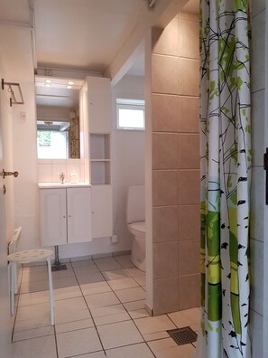 A bathroom with a toilet, sink, and cabinet. There is a chair and a window. - Pyttegaarden Apartments (Gudhjem)