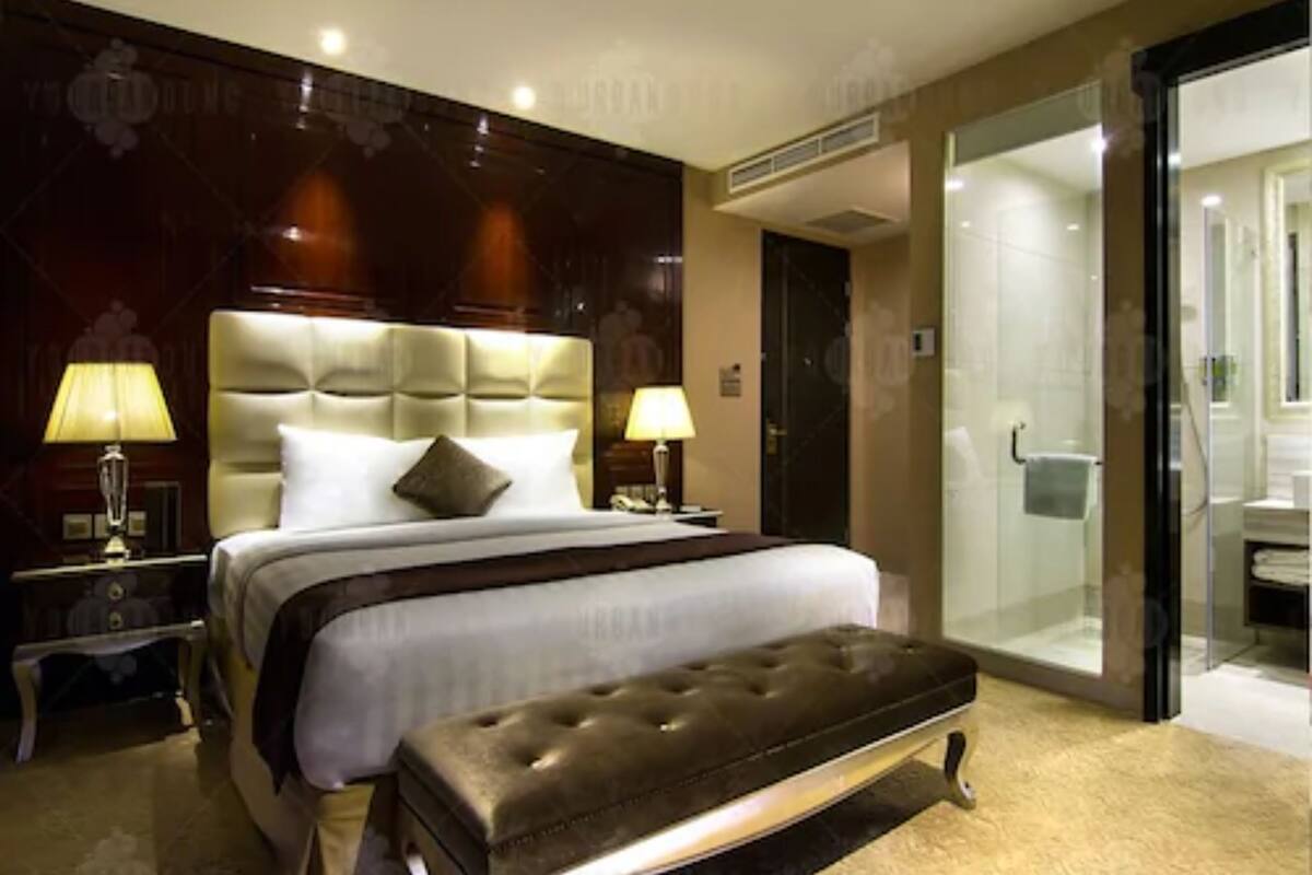 Habitación de hotel con una cama grande, dos mesitas de noche con lámparas, un banco de tonos oscuros y una zona de ducha con mampara de vidrio.