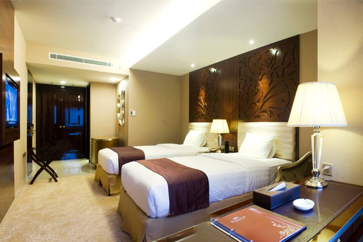 Habitación de hotel con dos camas, un escritorio con lámpara, un televisor y un panel decorativo en la pared.