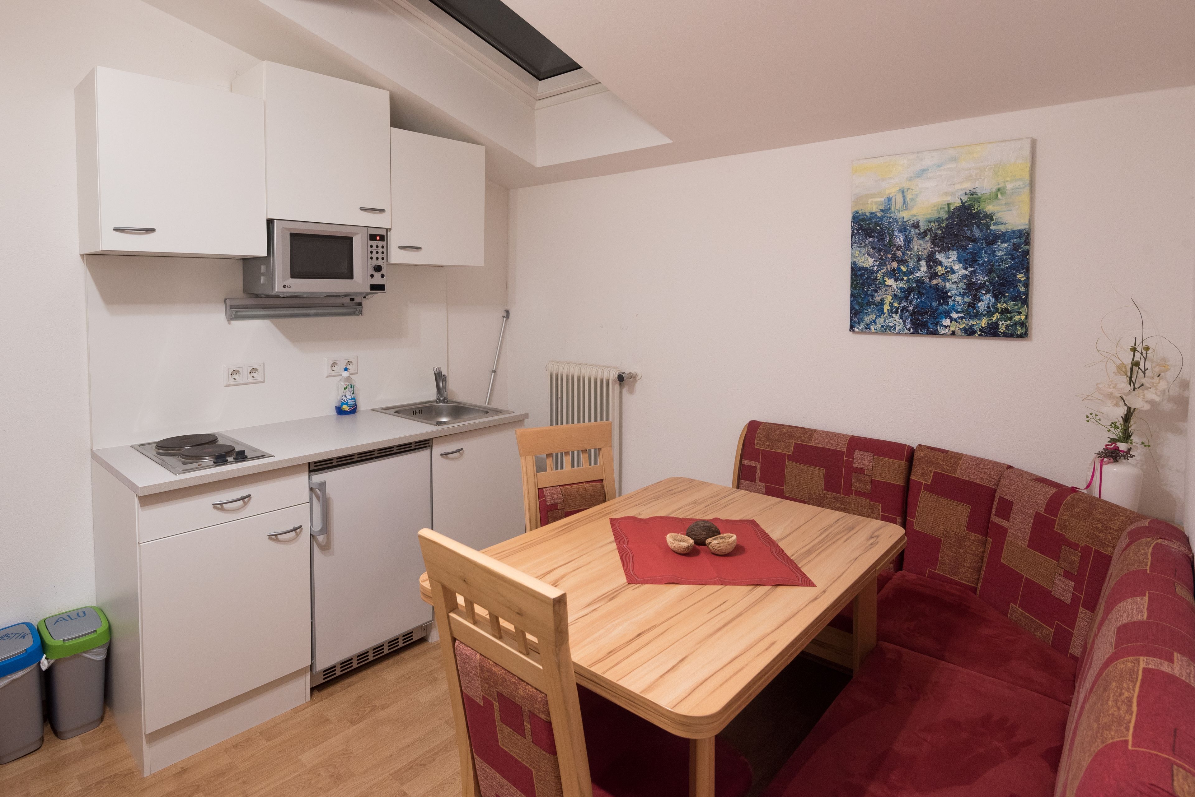 A compact kitchen with a microwave, sink, and cabinets. There is a dining table with chairs and a sofa. A painting is on the wall.