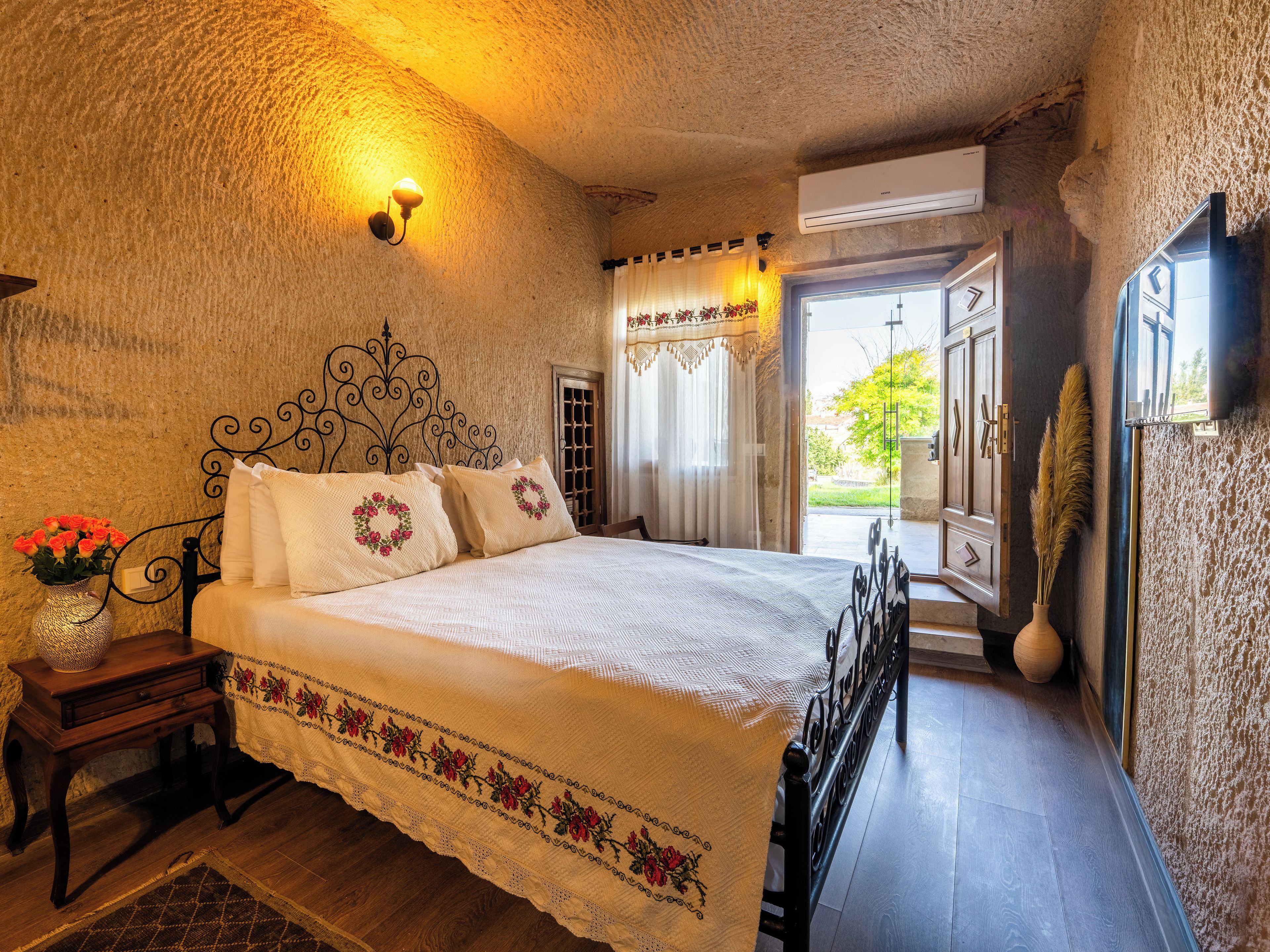 A bedroom with a bed, a nightstand, a vase with flowers, and a view of the outdoors.