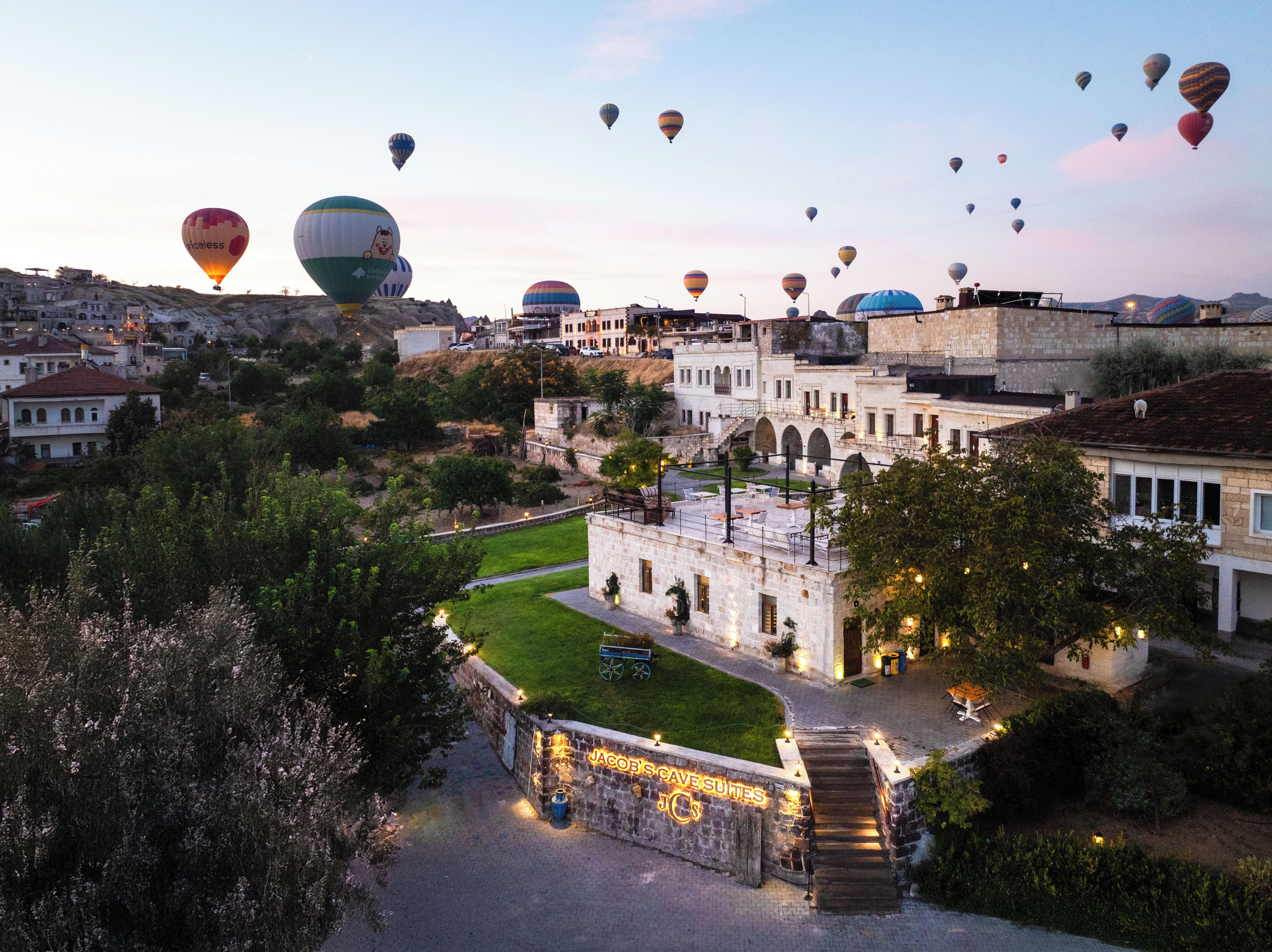 A historic building complex with a courtyard, surrounded by trees and a view of hot air balloons in the distance.