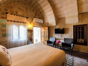 A room with a bed, two chairs, a television, and an arched stone ceiling. - Jacob's Cave Suites (Avanos)