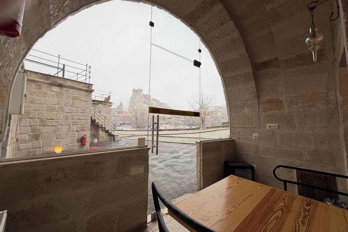 Habitación con paredes de piedra, una mesa de madera y vistas a un río y al paisaje urbano a través de un ventanal arqueado.