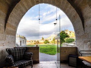 A room with a checkered sofa, a table, and a glass door leading to an outdoor area with a view of a hillside and buildings. - Jacob's Cave Suites (Avanos)