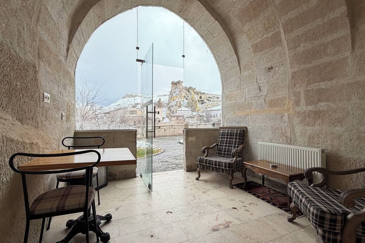Una habitación acogedora con una pared de piedra, una mesa de madera, dos sillas, un banco y vistas a un paisaje nevado a través de un amplio ventanal de arco.