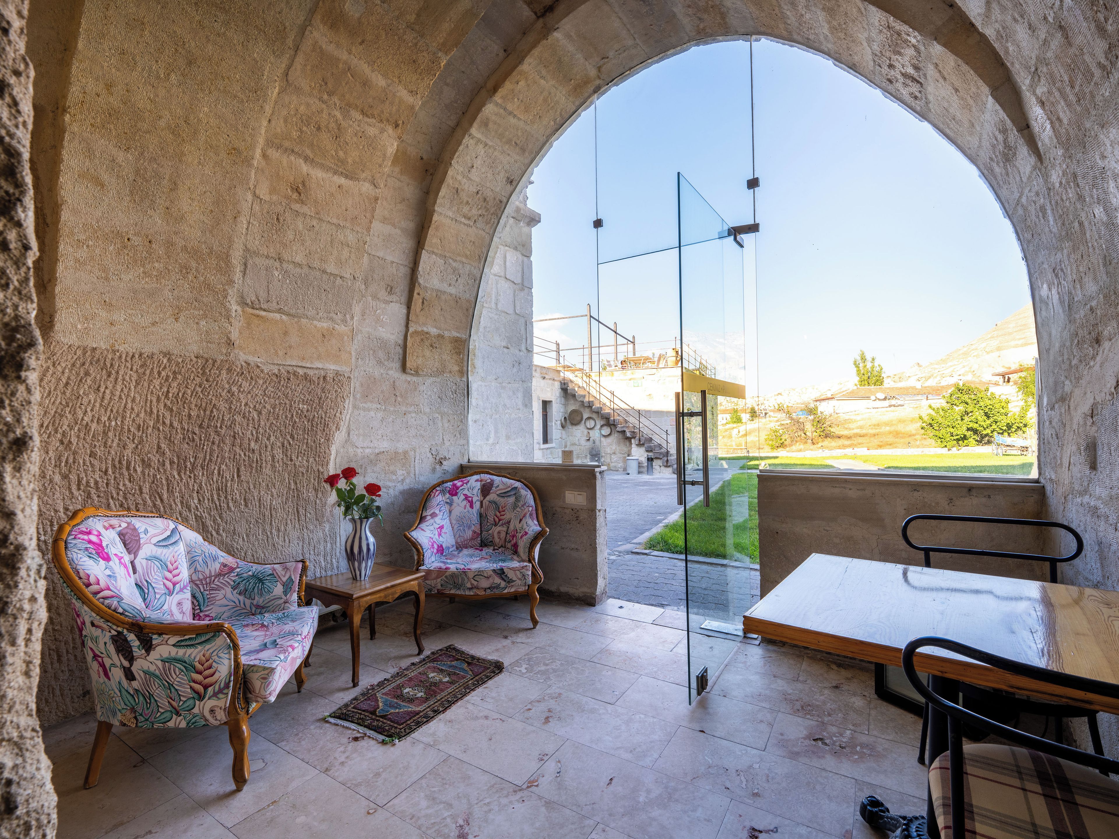 Una habitación de piedra con arco, con vistas a una colina, amueblada con sillas estampadas, una mesa y un jarrón con flores.
