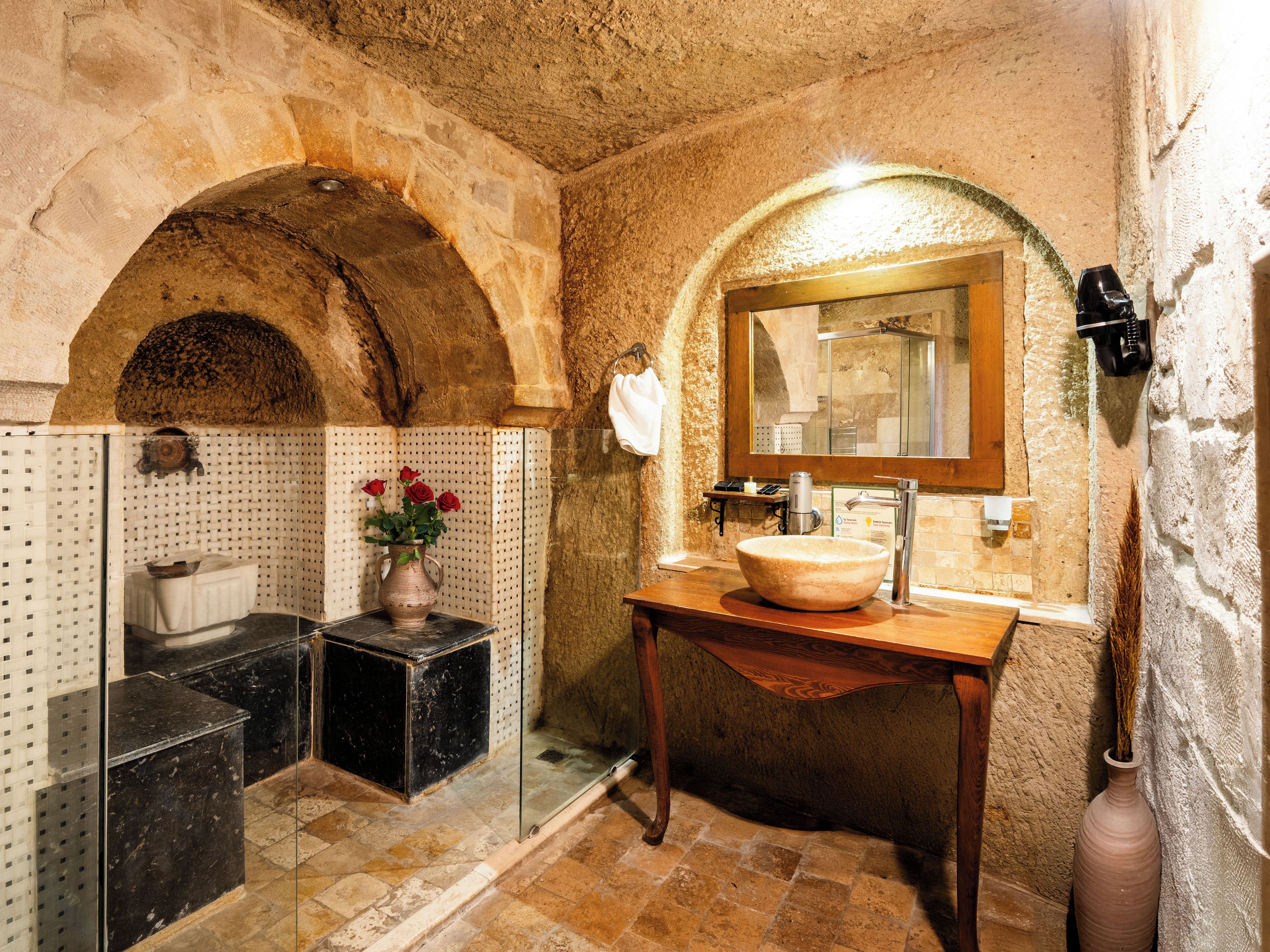 Un baño con pared de piedra, tocador de madera, un espejo grande y un jarrón con flores.