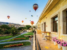 Un balcón con mobiliario exterior, un arreglo floral y vistas a un pueblo con globos aerostáticos a lo lejos.