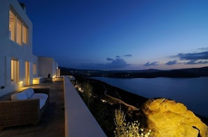A modern building with a balcony overlooking a calm body of water, featuring a seating area with cushions. - Villa Joy (Mykonos)