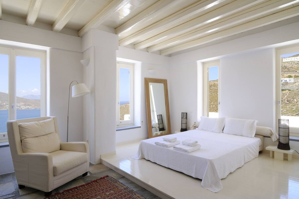 Un dormitorio con cama, una silla, un espejo y ventanas amplias.
