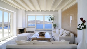 Un salon spacieux doté d’une grande fenêtre offrant une vue sur la mer et les montagnes. On y trouve un canapé blanc, une table basse et une cheminée.