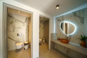 A bathroom with marble walls, a sink with a round mirror, and a toilet. - Nana Angela Apartments (Hersonissos)
