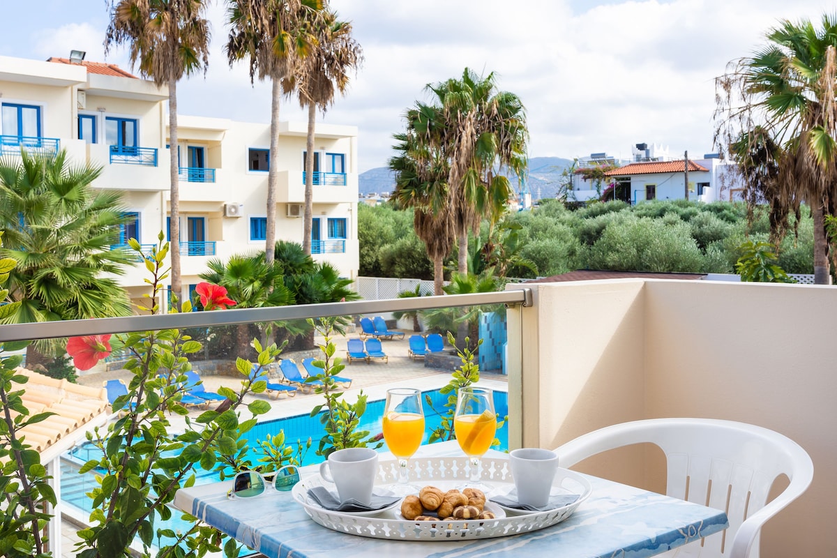 A balcony with a table set for breakfast, overlooking a swimming pool and a building with blue balconies.