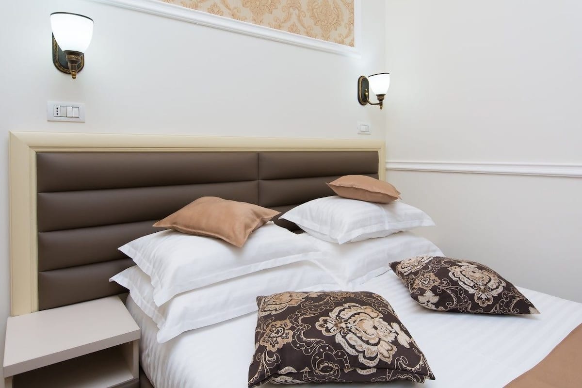 Una cama bien hecha con cabecera acolchada, dos lámparas fijadas a la pared y una mesita de noche pequeña.