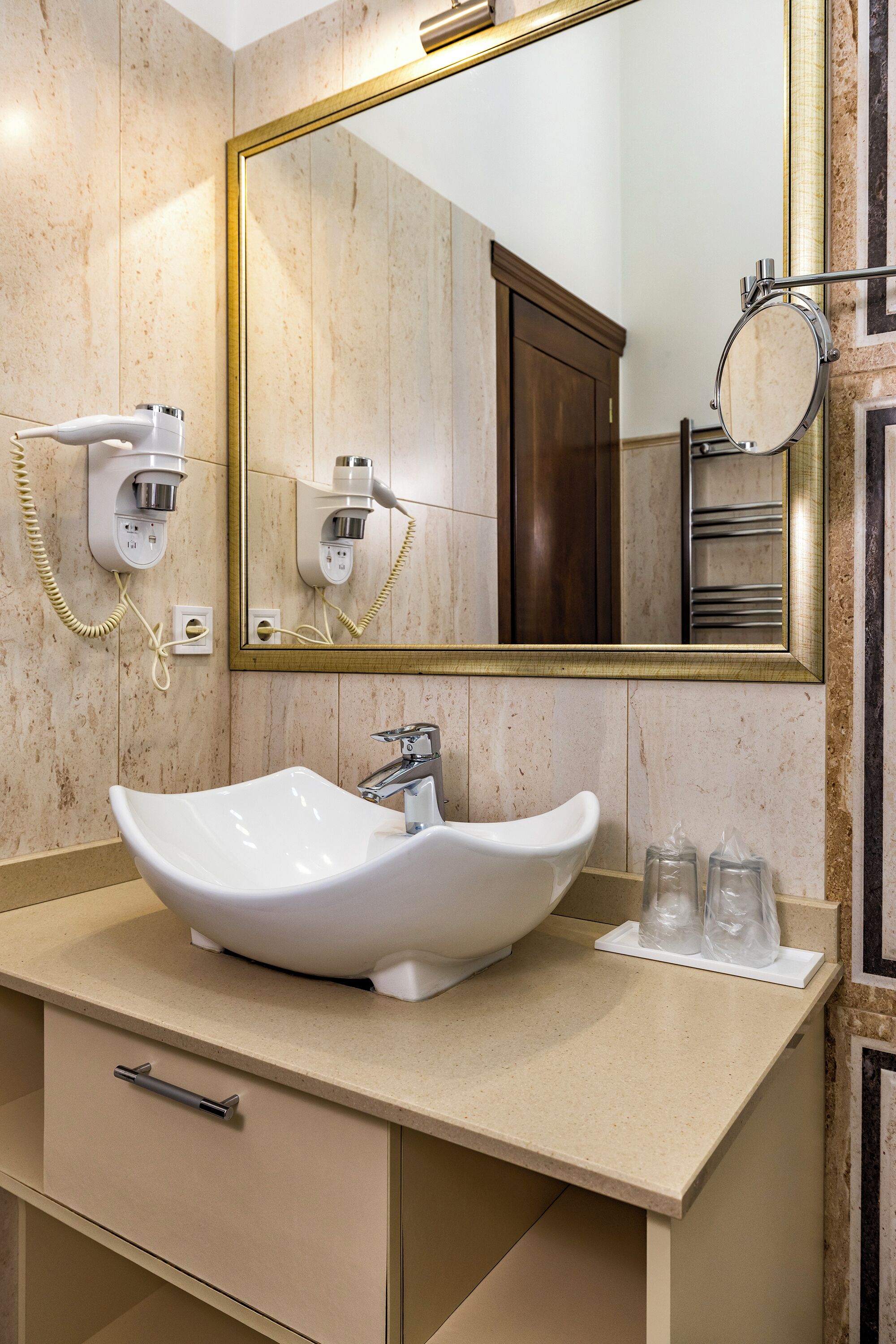 A modern bathroom with a beige marble wall, a large mirror, a white vessel sink, and a hairdryer mounted on the wall.
