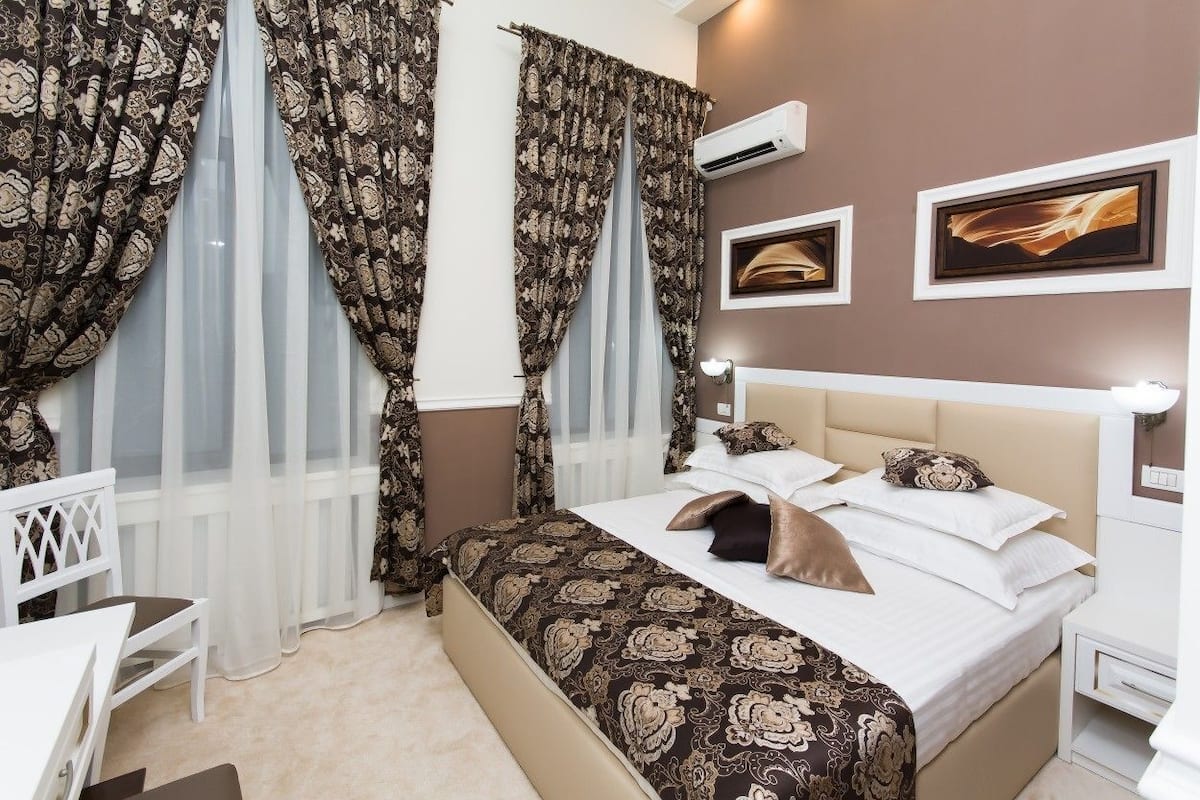 Un dormitorio con cama, cortinas, una mesita de noche, una silla y una obra de arte en la pared.