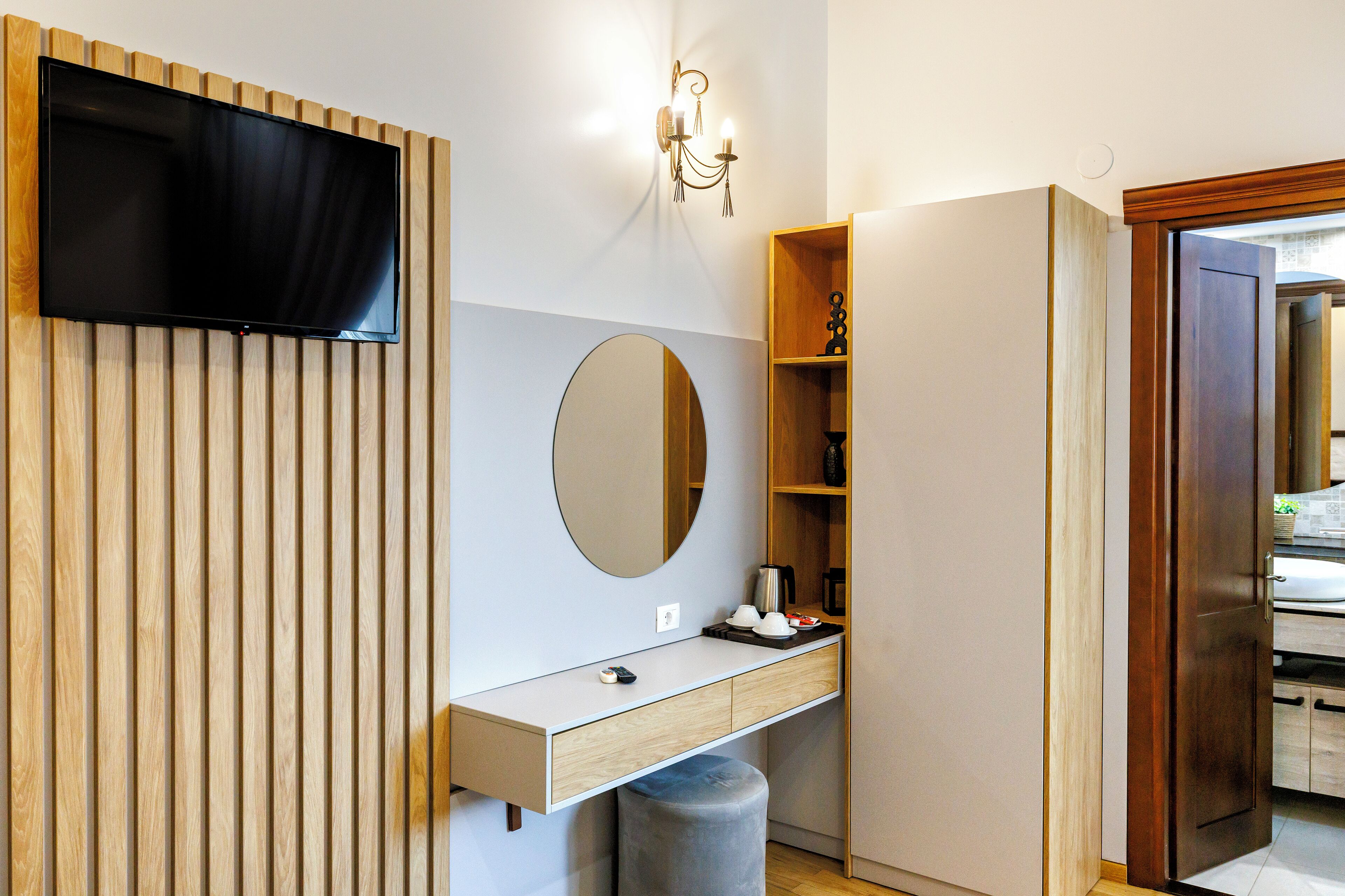 Un baño moderno con tocador de madera, espejo redondo, televisor montado y un estante pequeño con artículos de aseo.