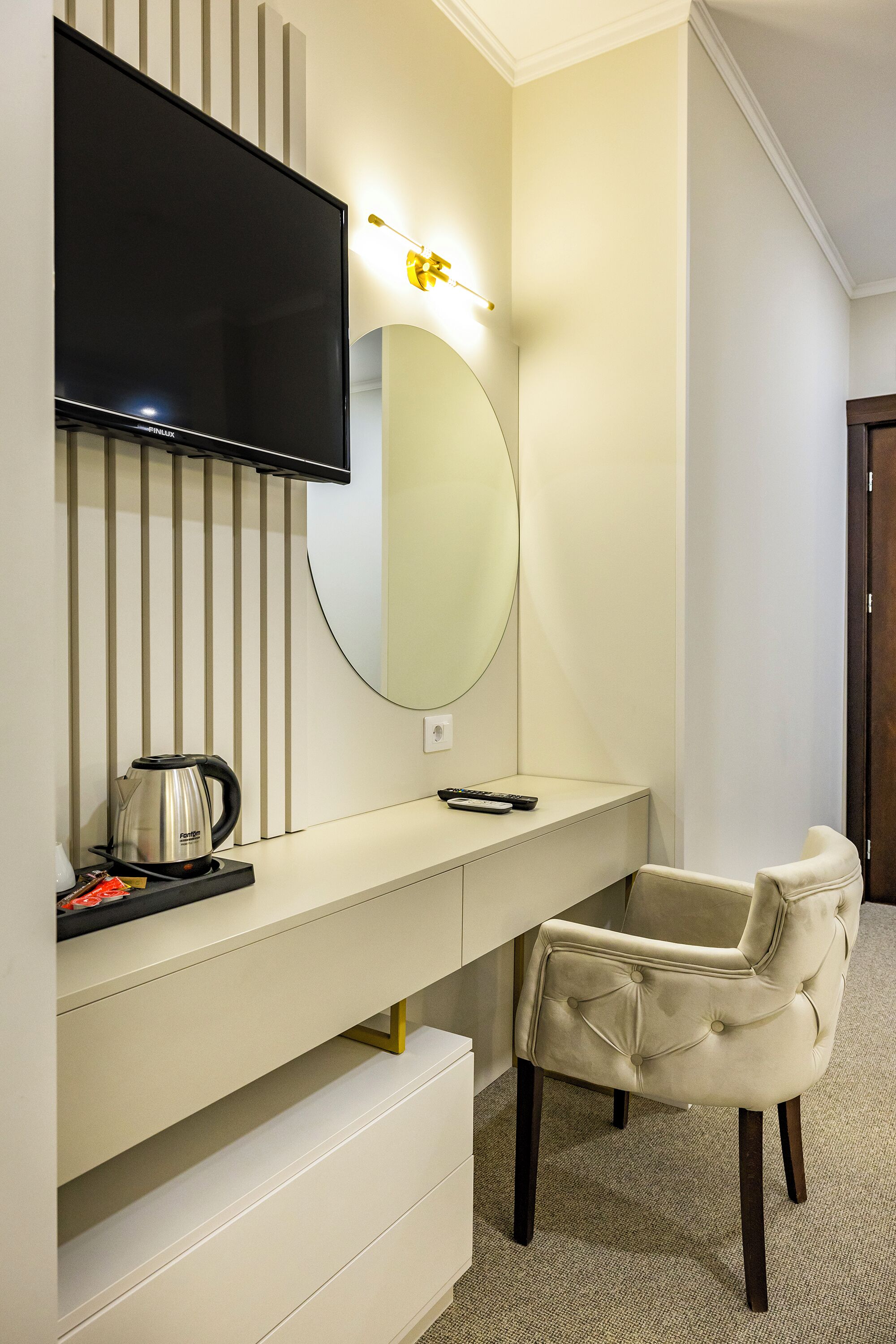 A modern hotel room with a flat-screen TV mounted on the wall, a large mirror, a beige upholstered chair, a white console table with a kettle and remote control, and a carpeted floor.