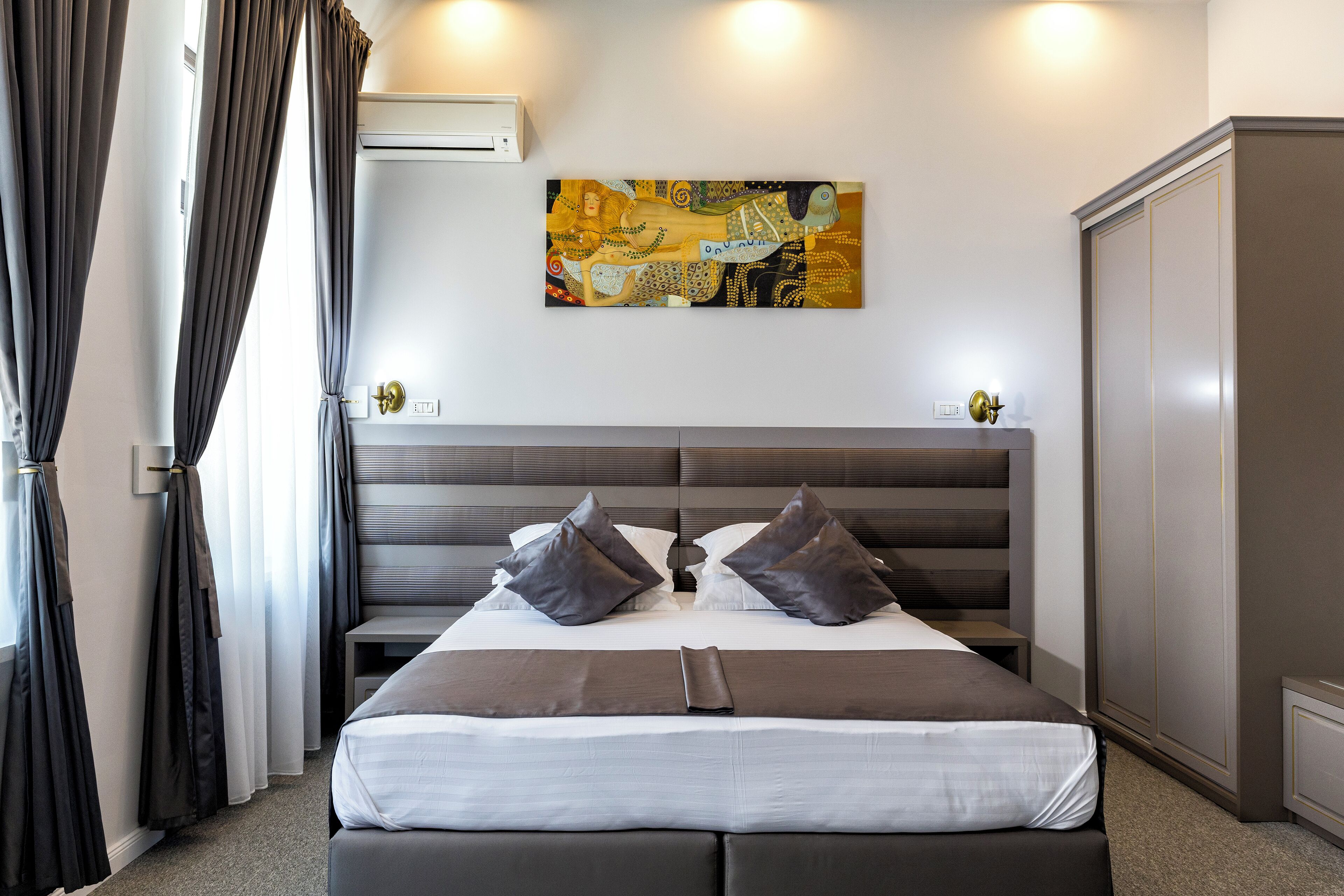 A modern hotel room with a large bed, dark curtains, a headboard with integrated lighting, and a wall-mounted artwork.