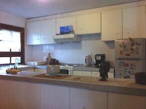 Eine Küche mit weißen Schränken, einem Fenster mit Vorhängen, einem Wasserkocher, einer Kaffeemaschine und einem Kühlschrank mit Magneten.