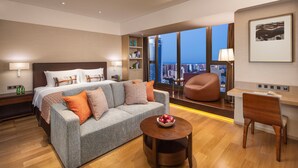 Una habitación de hotel moderna con una cama grande, un sofá gris, una mesa de centro de madera y un escritorio con una silla. La habitación tiene un ventanal con vistas a la ciudad.