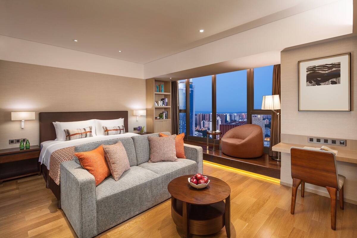 Een moderne hotelkamer met een groot bed, een grijze bank, een houten salontafel en een bureau met een stoel. De kamer heeft een groot raam met uitzicht op de stad.