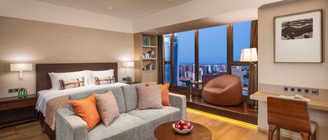 Una habitación de hotel moderna con una cama grande, un sofá gris, una mesa de centro de madera y un escritorio con una silla. La habitación tiene un ventanal con vistas a la ciudad.