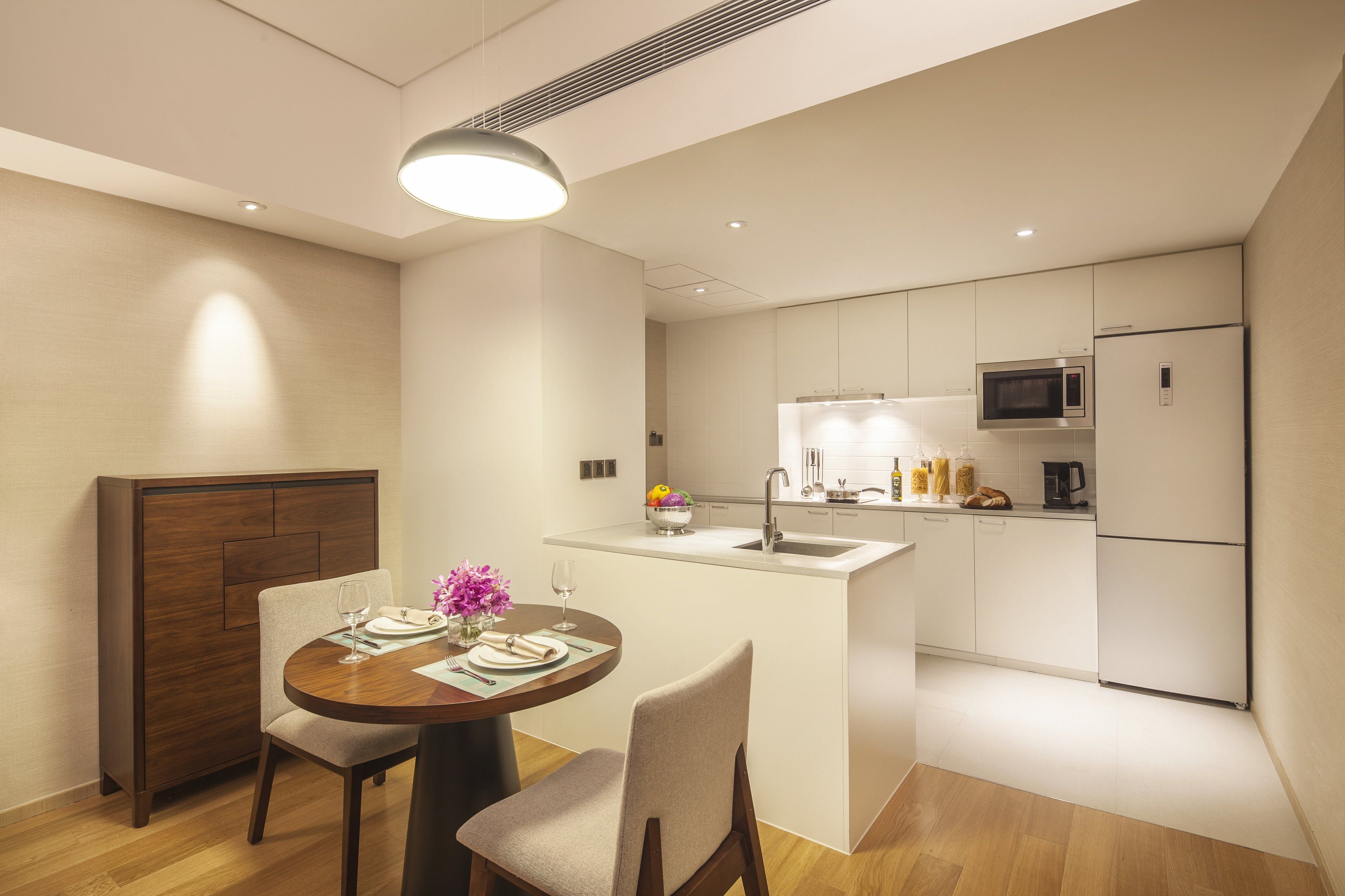 A modern kitchen with white cabinets, a microwave, and a refrigerator. There is a dining area with a wooden table and chairs, and a small island with a sink.