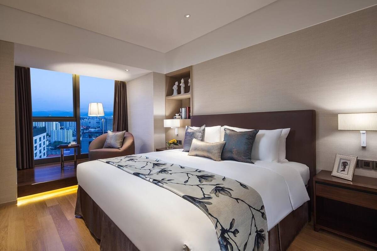 Een moderne hotelkamer met een groot bed, uitzicht over de stad, een zithoek met een stoel en een nachtkastje met een lamp.