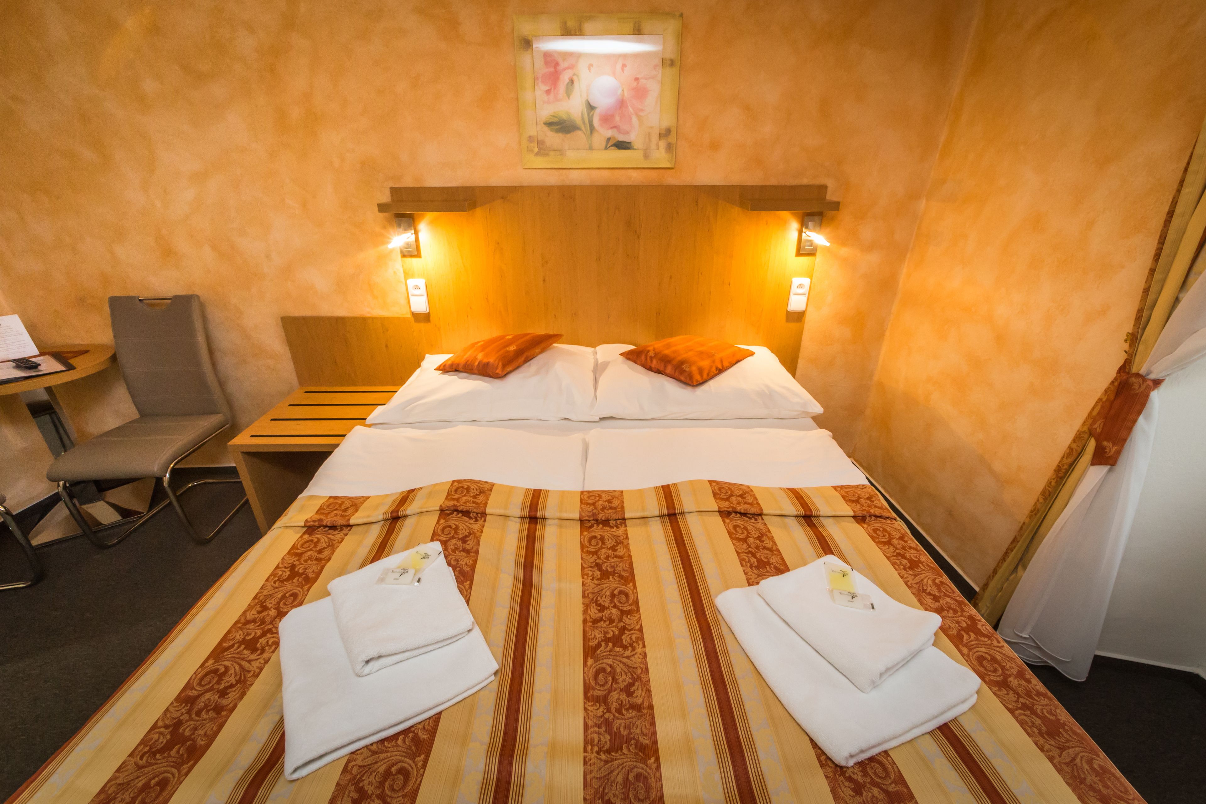 A hotel room with a bed, two pillows, a bedside table, a chair, and a painting on the wall.