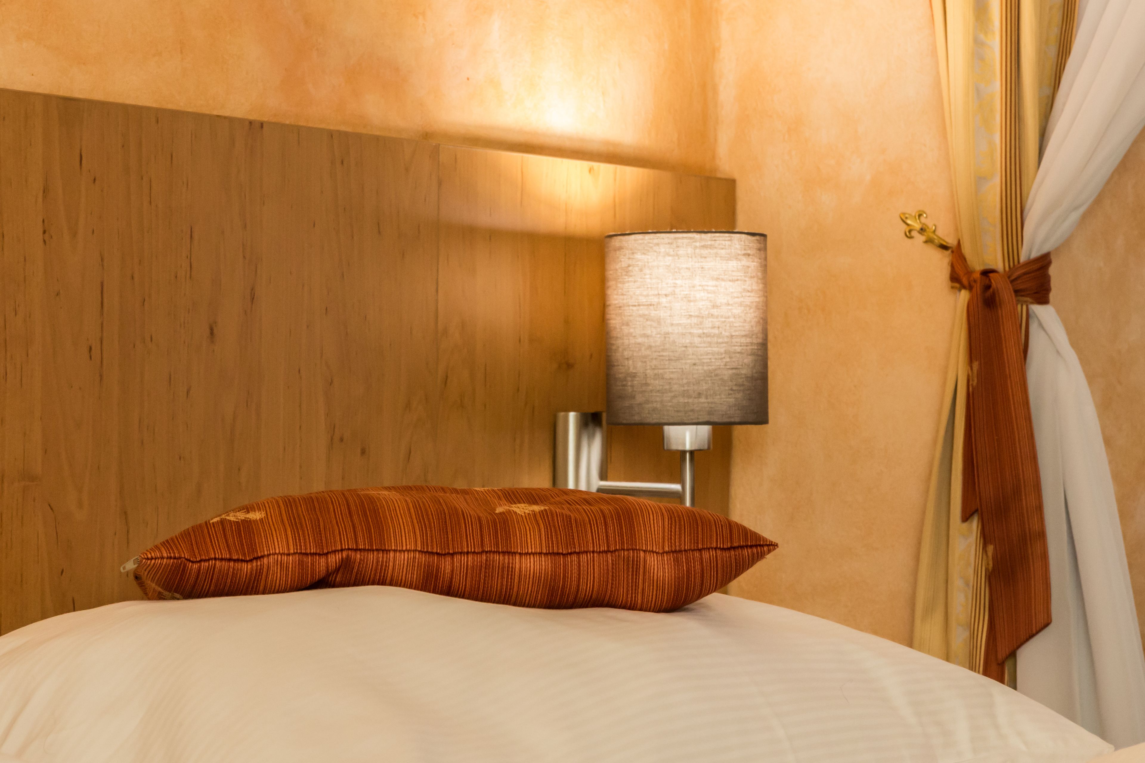 A hotel room with a wooden headboard, a white bed with a striped pillow, a bedside lamp, and a curtain tied back with a brown ribbon.