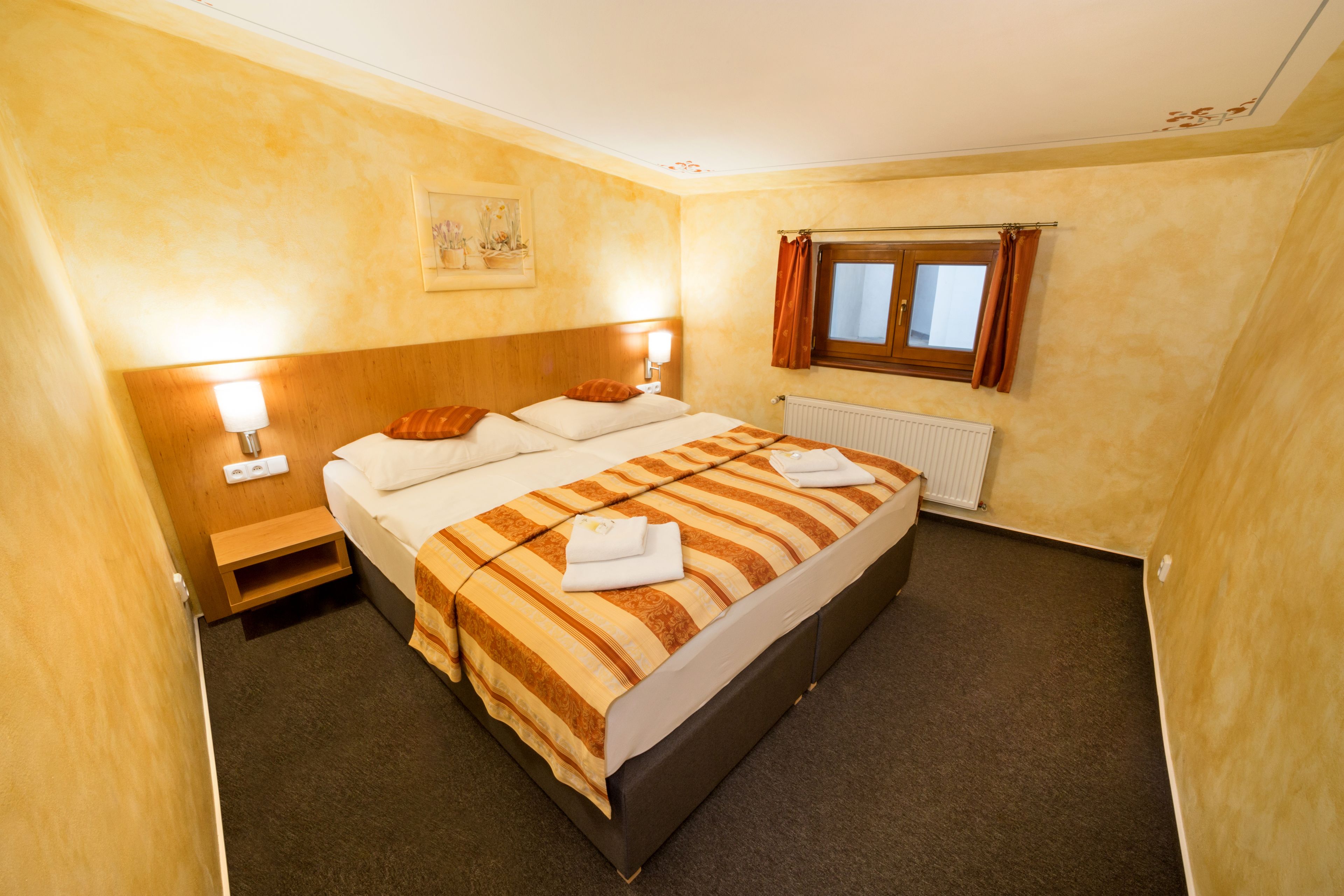 A hotel room with a large bed, two bedside tables, a window with curtains, a radiator, and a wall-mounted artwork.