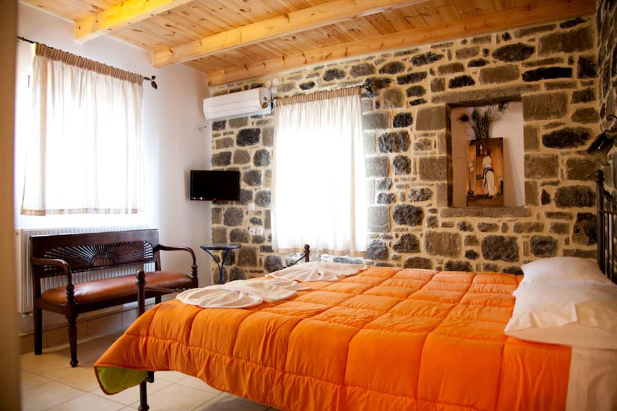 Un dormitorio con pared de piedra, techo de madera, cama con colcha naranja, un banco y televisión en la pared.