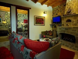A cozy living room with a stone fireplace, a sofa with floral cushions, a television mounted above the fireplace, and a framed painting on the wall.