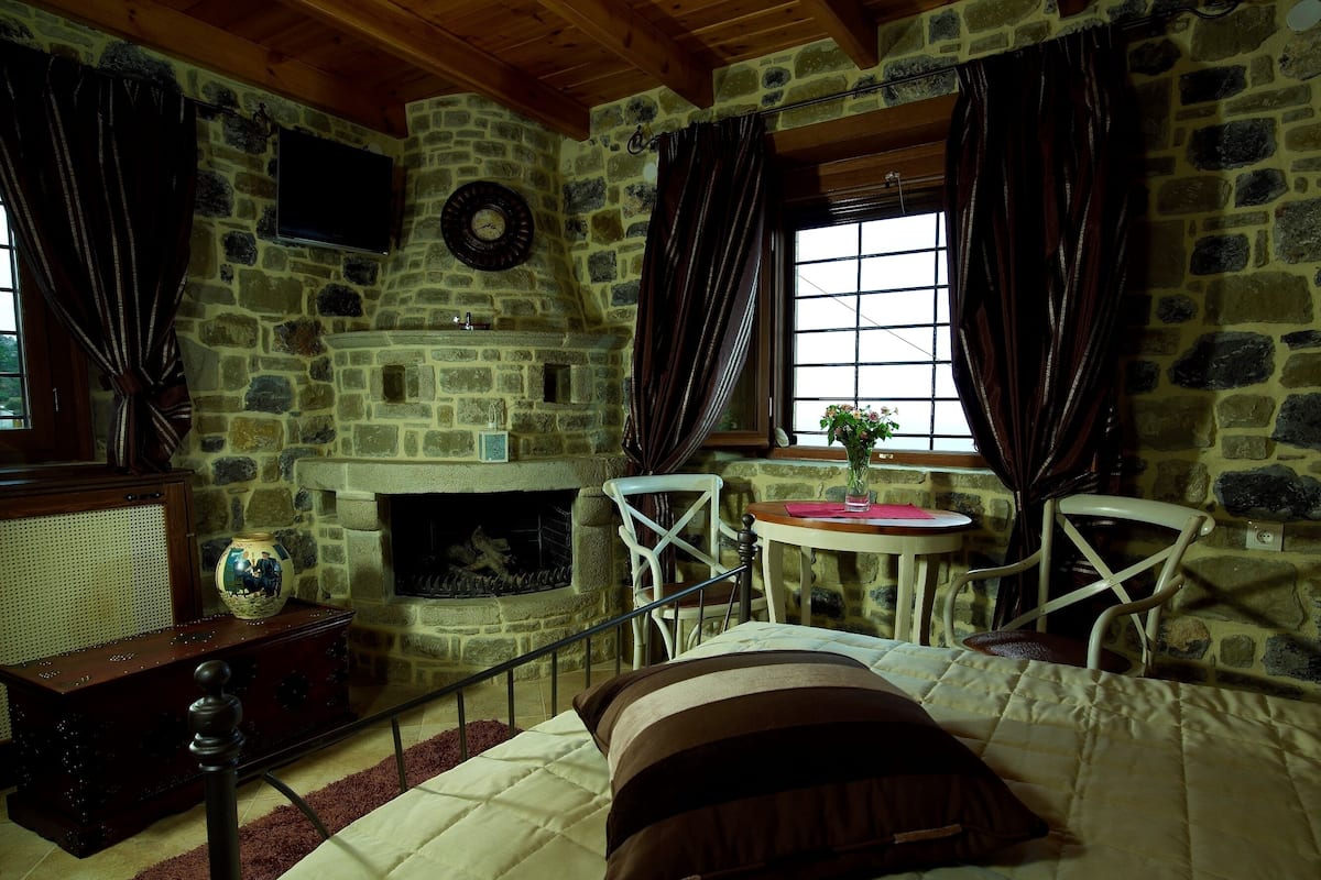 Una habitación de paredes de piedra con chimenea, una cama, una mesa con un florero y dos sillones.